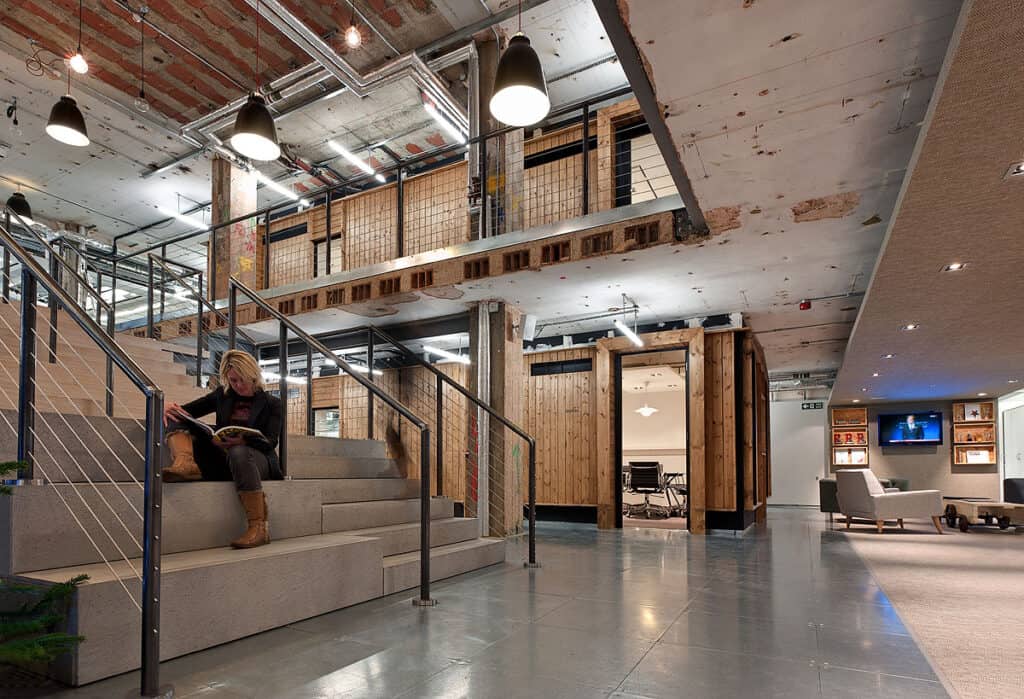 Fit Out Guide Refurbished industrial-style office featuring exposed ceilings, ductwork, and wooden paneling. A person sits reading on a staircase with metal railings, while the background showcases glass-walled rooms with wooden frames and an open seating area with sofas and chairs under hanging ceiling lamps.