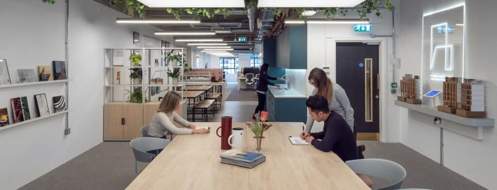 about-TEMP Refurbished open-plan office featuring a large wooden table with professionals collaborating on documents, surrounded by shelves with plants and books, a kitchenette area, and an open workspace. The ceiling showcases exposed pipes and hanging greenery, creating a modern and eco-friendly work environment.