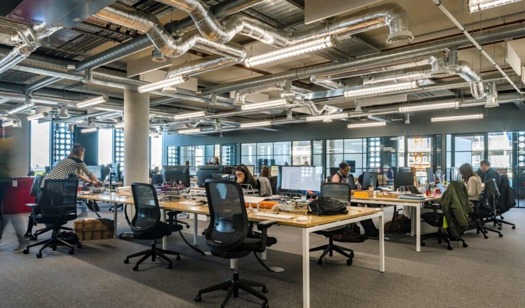 slider-3 Refurbished open-plan office with rows of desks equipped with monitors, chairs, and office supplies. The space features exposed ductwork, industrial lighting, and large windows that flood the room with natural light, creating a productive and modern work environment.