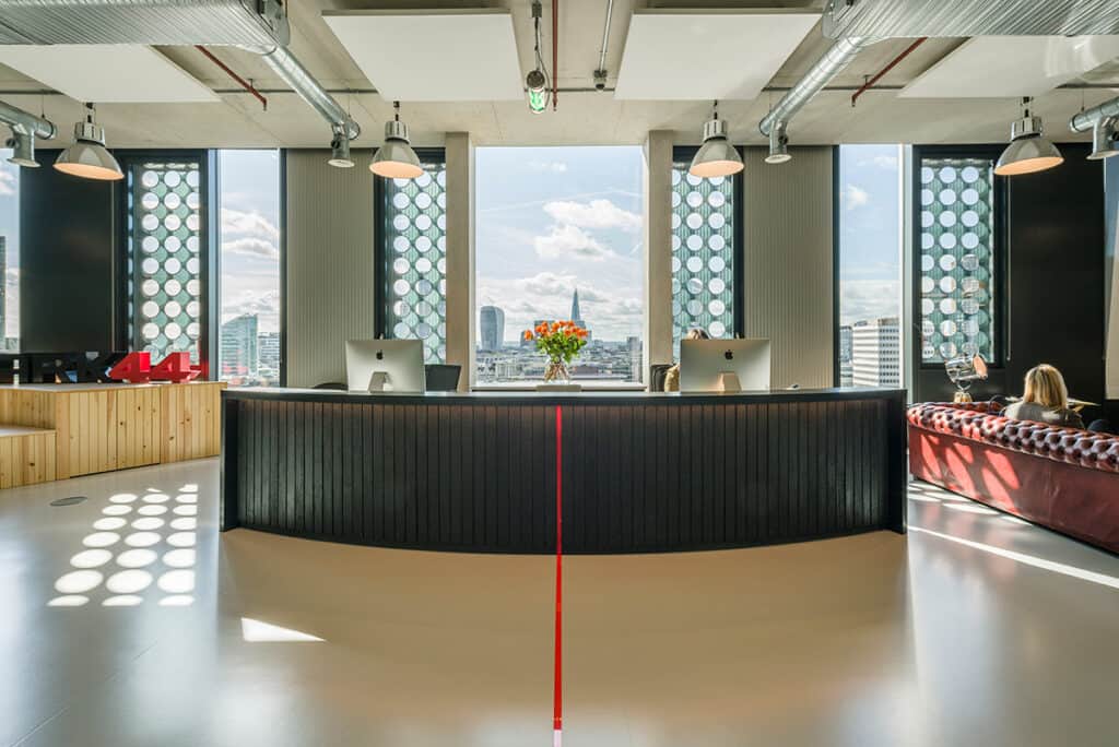 _1 Refurbished office reception area with a sleek black curved desk, dual iMacs, and vibrant orange floral arrangement. Tall circular-patterned windows offer views of London landmarks including the Shard and Walkie Talkie building. Red leather seating and industrial ceiling design complete the modern, welcoming aesthetic.
