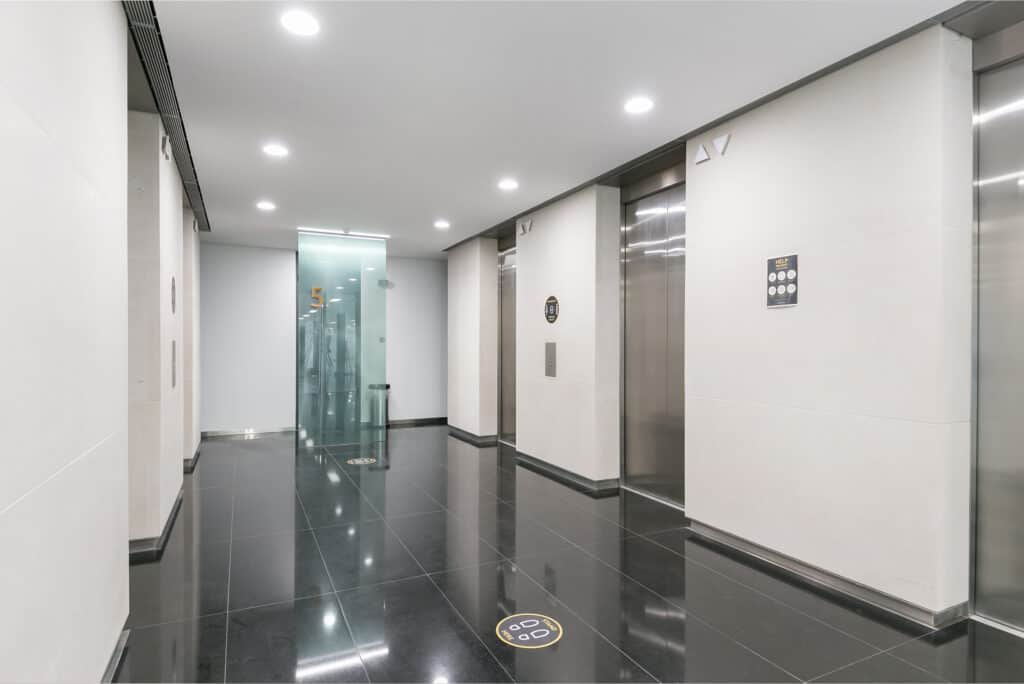 100-wood-street-3 A modern elevator lobby with three elevator doors, each featuring an indicator light showing direction. The floor is covered in shiny black tiles, and the white walls contrast with the sleek design. A glass wall on the left displays the number "5" for the floor level. Recessed ceiling lights provide bright illumination.