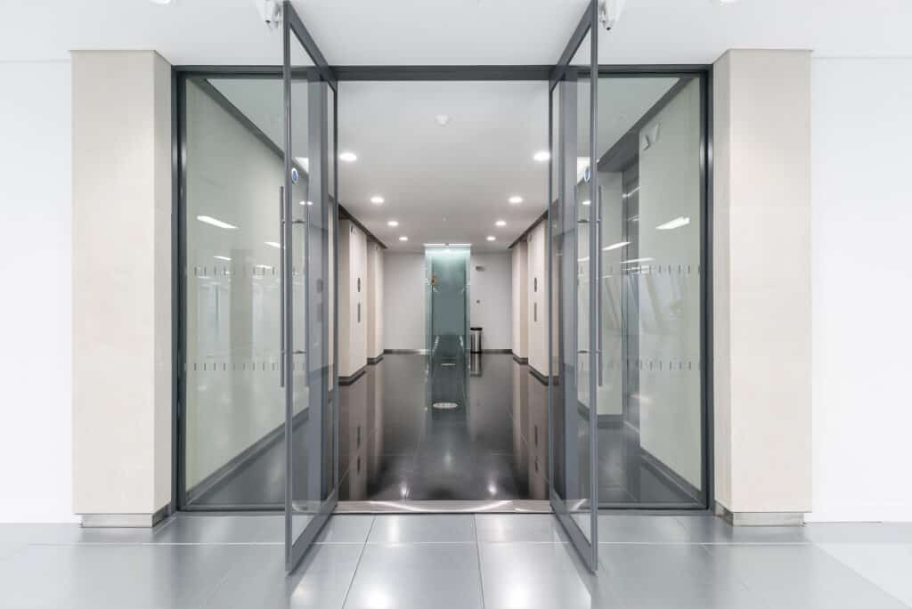 100-wood-street-4 A modern, clean hallway with polished reflective flooring and glass walls on both sides. Ceiling lights provide bright illumination. Glass doors mark both the entrance and the far end of the hallway, contributing to the sleek, minimalistic design.