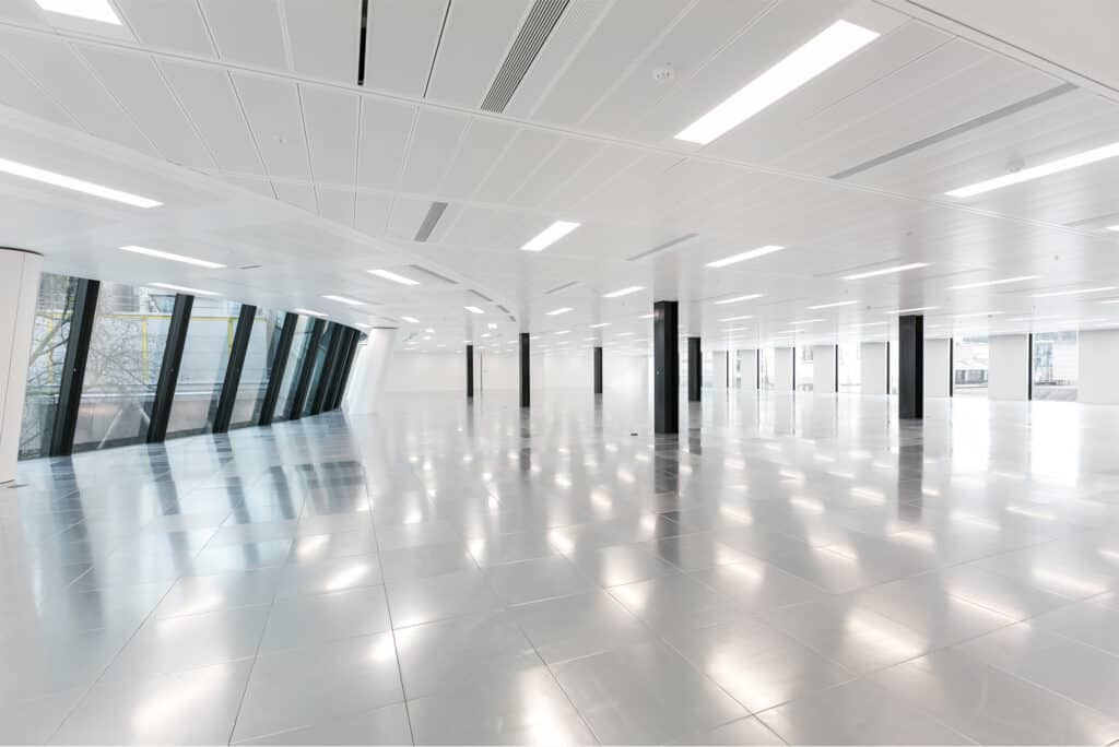 Building Refurbishment A large, modern office space with a highly reflective floor, numerous overhead lights, and large, slightly tilted windows on the left side allowing natural light in. Black columns are evenly spaced throughout the room, contributing to the architectural design.