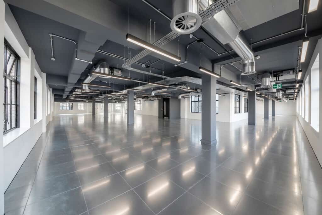 M&E Contractor London A large, modern industrial-style interior with polished reflective floors and exposed ceiling ducts and pipes. Natural light streams in through large windows on both sides. Structural columns are spaced throughout, with overhead lighting fixtures and an emergency exit sign visible in the background.