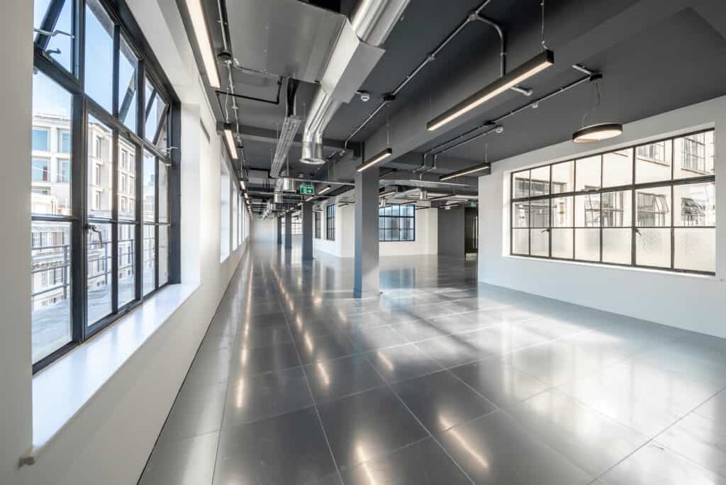 11-jewry-street-3 A modern, empty office space with polished tile flooring and large windows on both sides, allowing natural light to fill the room. The ceiling features exposed ductwork and lighting fixtures, with structural columns spaced throughout. An emergency exit sign is visible in the distance.