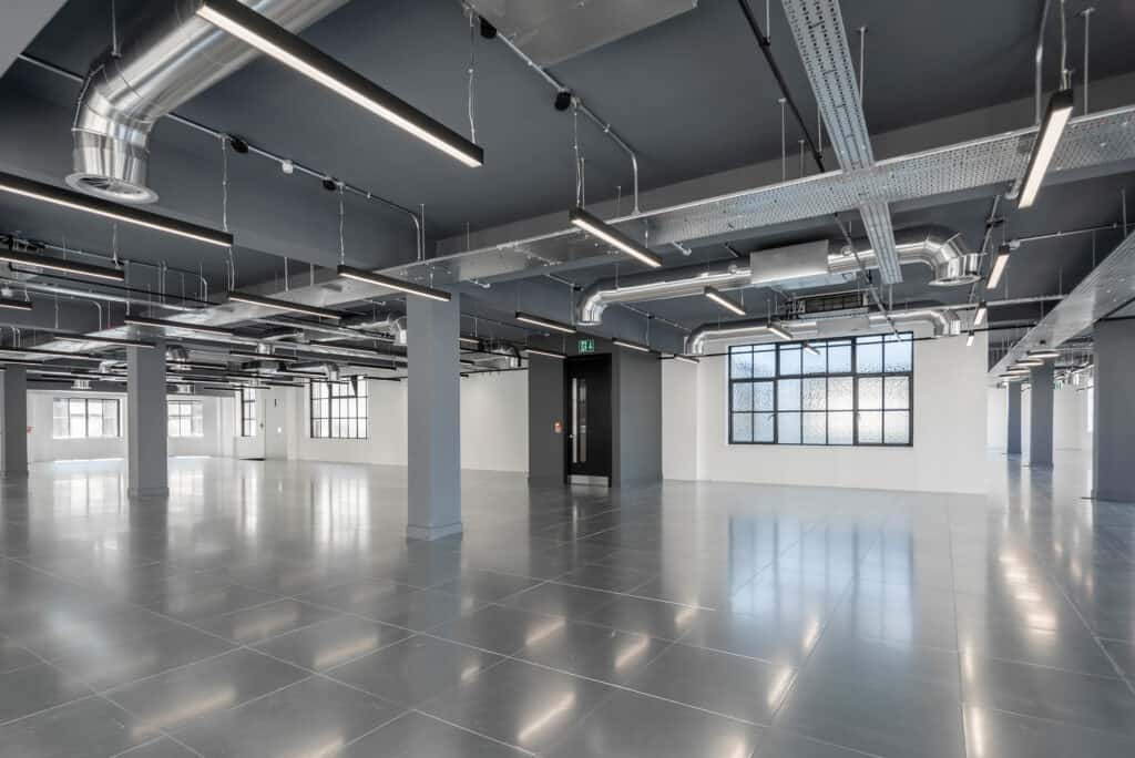 11-jewry-street-5 A large, CAT A modern room with an industrial design featuring exposed ductwork and piping on the ceiling. The polished tile floor reflects the natural light coming through large windows. Structural columns are spaced throughout the space, and a black door is centered on one wall. Linear light fixtures provide overhead illumination.
