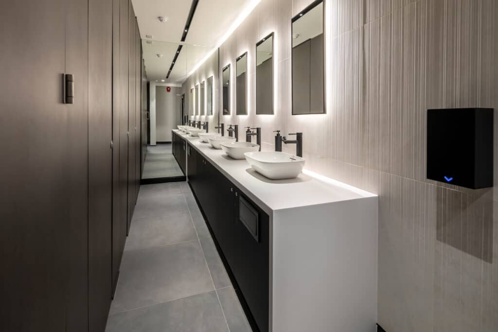 11-jewry-street-6 A modern public restroom with a long white countertop featuring multiple vessel sinks and black faucets. Rectangular mirrors with backlighting hang above the sinks. The gray tiled floor complements the dark-colored stall doors on the left and a black hand dryer mounted on the right wall, creating a sleek, contemporary design.