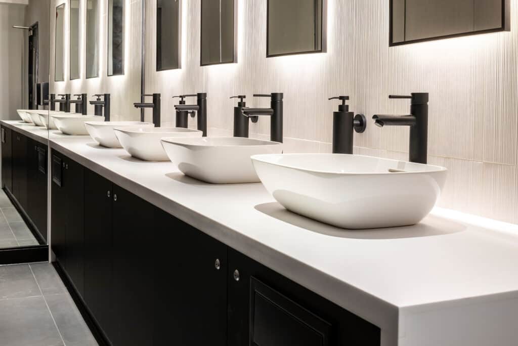 11-jewry-street-7 A modern bathroom featuring five white vessel sinks mounted on a long countertop, each with a black taps. Mirrors are positioned above each sink, and bright, even lighting enhances the clean, minimalist design.