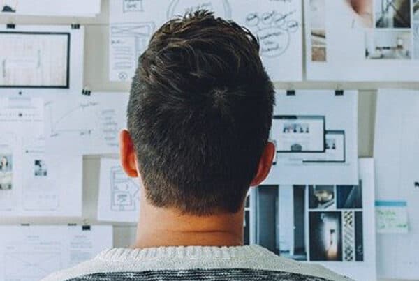 Back view of a person looking at a wall covered with papers, diagrams, sketches, charts, and photographs, suggesting design or planning work.