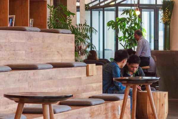 A modern, open interior space featuring wooden seating areas and tables. In the foreground, two people are engaged in conversation, while others are seen in the background, creating a relaxed and social atmosphere. Green plants add a touch of nature to the setting.
