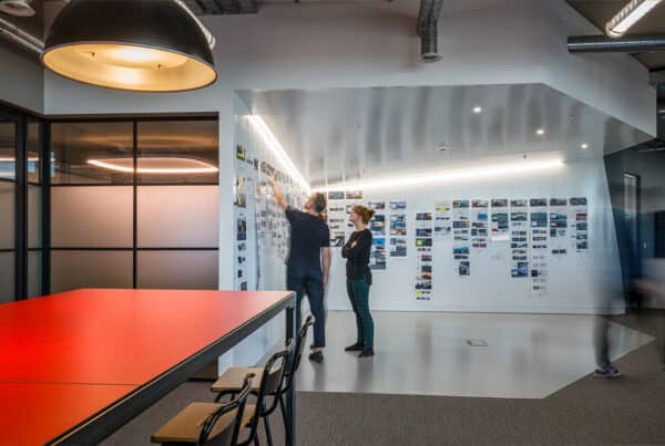 Modern office space with two people standing in front of a wall covered in images and documents. One person points at the wall while the other observes with arms crossed. A large red table with chairs is in the foreground, and glass partitions are on the left. The room is brightly lit with overhead lighting.