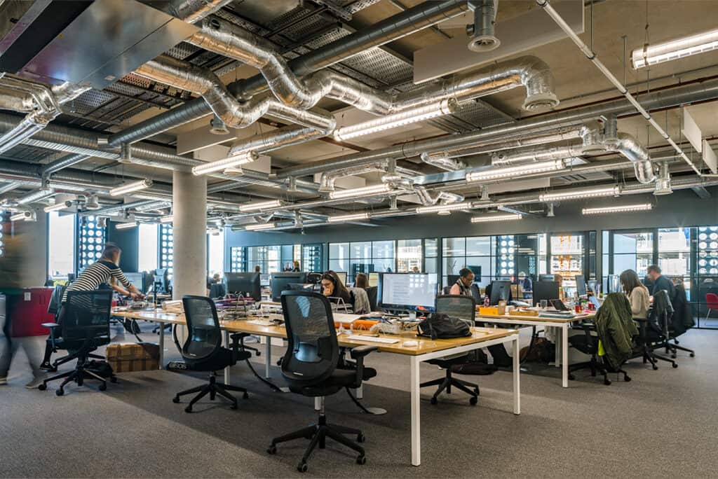 _9 Refurbished open-plan office with rows of desks equipped with monitors and ergonomic chairs. Industrial ceiling design with exposed ductwork and pendant lighting complements large rear-facing windows that fill the space with natural light. A modern, functional workspace designed for productivity and collaboration.