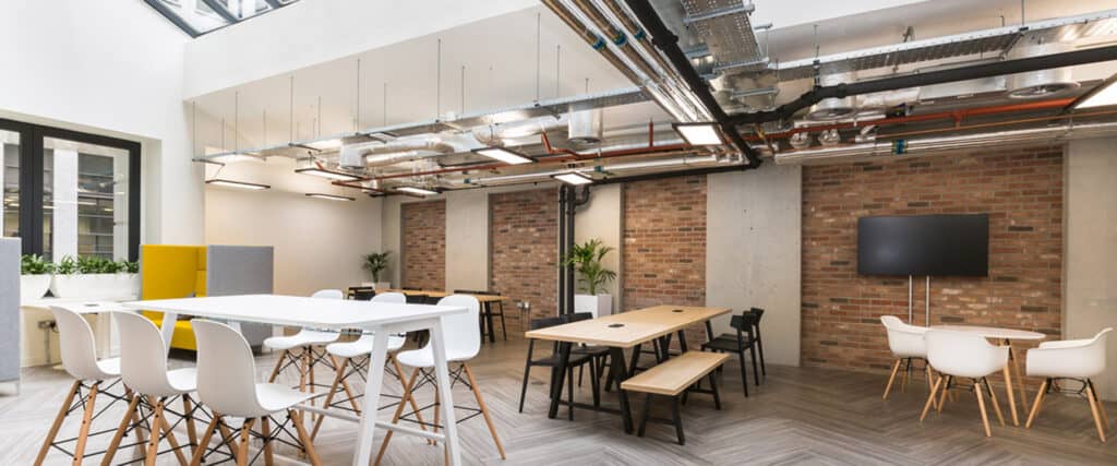 How-to-attract-new-tenants-during-a-pandemic Modern industrial-style office space with high tables and white chairs, lower tables with benches, and exposed ceiling pipes and ducts. The room features brick and concrete walls, a large flat-screen TV mounted on a brick wall, and decorative plants throughout.