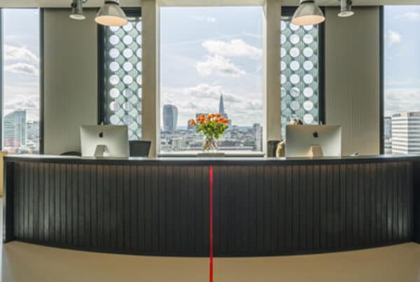 Refurbished office reception with modern curved desk, Apple iMacs, vibrant orange flowers, and city view through designer windows—showcasing contemporary workplace design.