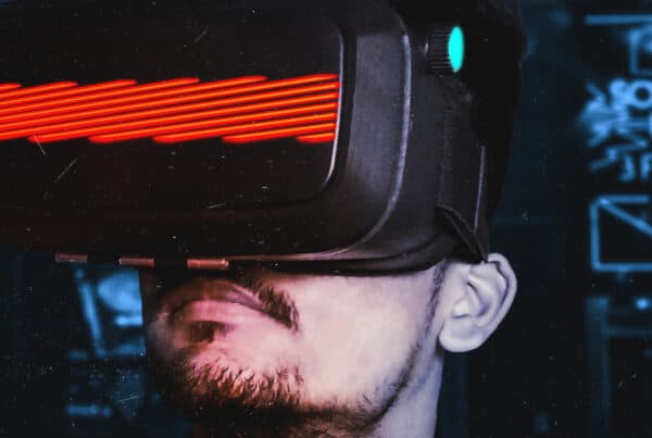 A person wearing a virtual reality headset, with a focus on their face. The headset features glowing red accents and is set against a blurred background that suggests a vibrant, tech-focused environment.