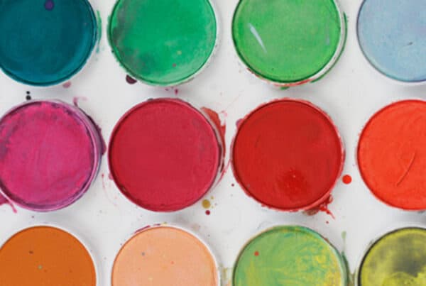 A vibrant array of circular paint pots arranged in rows, showcasing a variety of colors including reds, pinks, greens, and blues. The colorful paints have varying textures and some have been mixed, creating an artistic display.