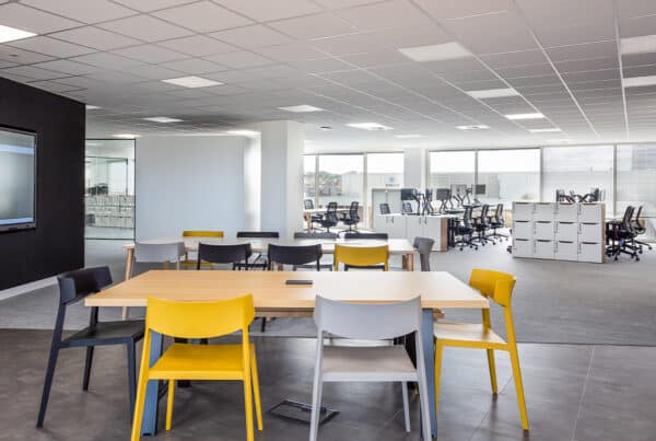 Modern open-plan CAT B office space with a casual seating area in the foreground featuring yellow, black, and gray chairs around tables. In the background, rows of desks with office chairs and computer monitors are visible. The space includes large windows for natural light, a black wall with a mounted screen, and a grid-pattern ceiling with recessed lighting.