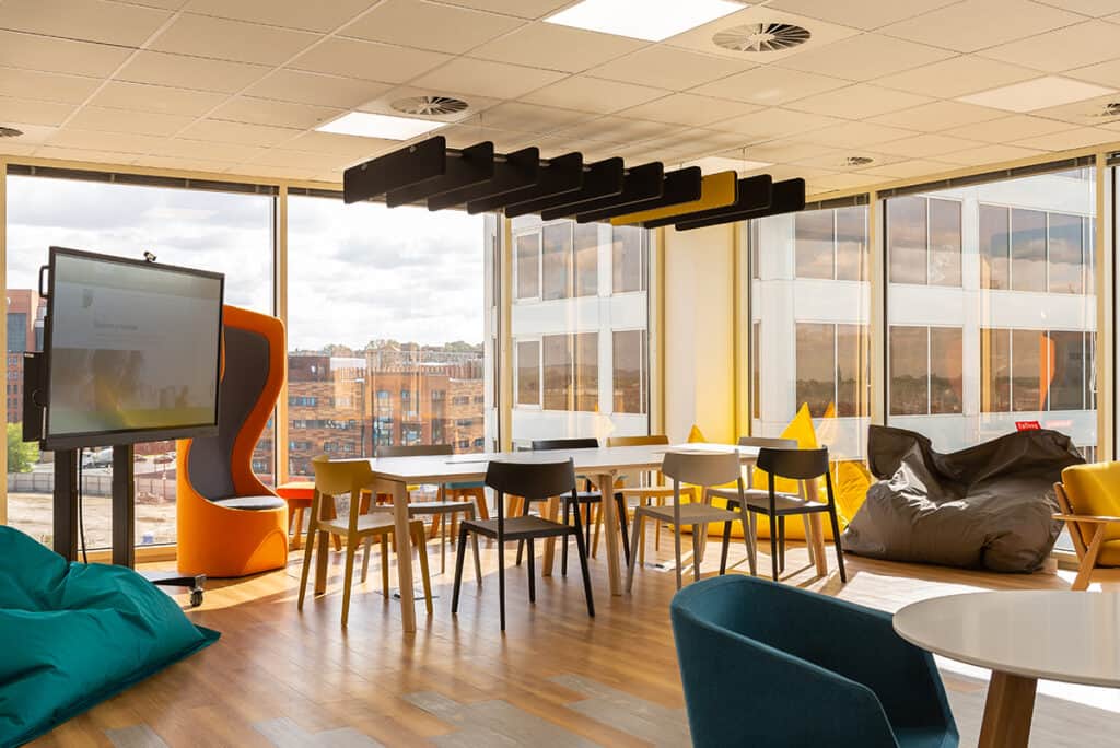 bcs-6 Modern office lounge area with large windows offering views of nearby buildings. Furnishings include tables with chairs, bean bags, an orange pod chair, and a large screen on a stand. The space is bright and filled with natural light.
