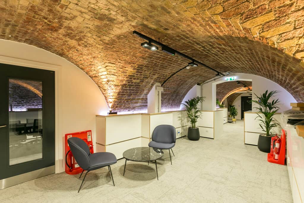 dolby_11 Office space with a vaulted brick ceiling, featuring two gray chairs and a small round glass table in the foreground. The area is well-lit with track lighting and decorated with several potted plants along the corridor. Fire safety equipment, including a fire extinguisher and hose reel, is visible near the entrance door on the left.