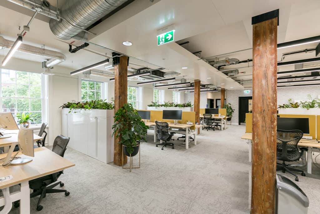 dolby_4 Refurbished office space with rows of modern workstations, large windows for natural light, and green plants enhancing the workspace ambiance. Industrial ceiling design with exposed ducts and wooden beams adds character, while white cabinets and emergency signage reflect a functional, refreshed layout.