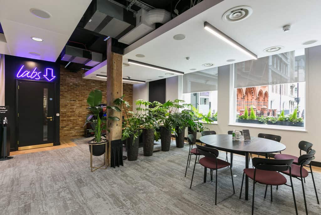 dolby_9 Modern office space featuring a mix of natural and industrial elements. A neon sign reading 'labs' with a downward arrow is above a door on the left. Large potted plants are arranged near the windows and in the center. On the right, a round table with chairs suggests a collaborative area. The space includes grey carpet flooring, exposed brick walls, wooden beams, and large windows with views of an urban environment.