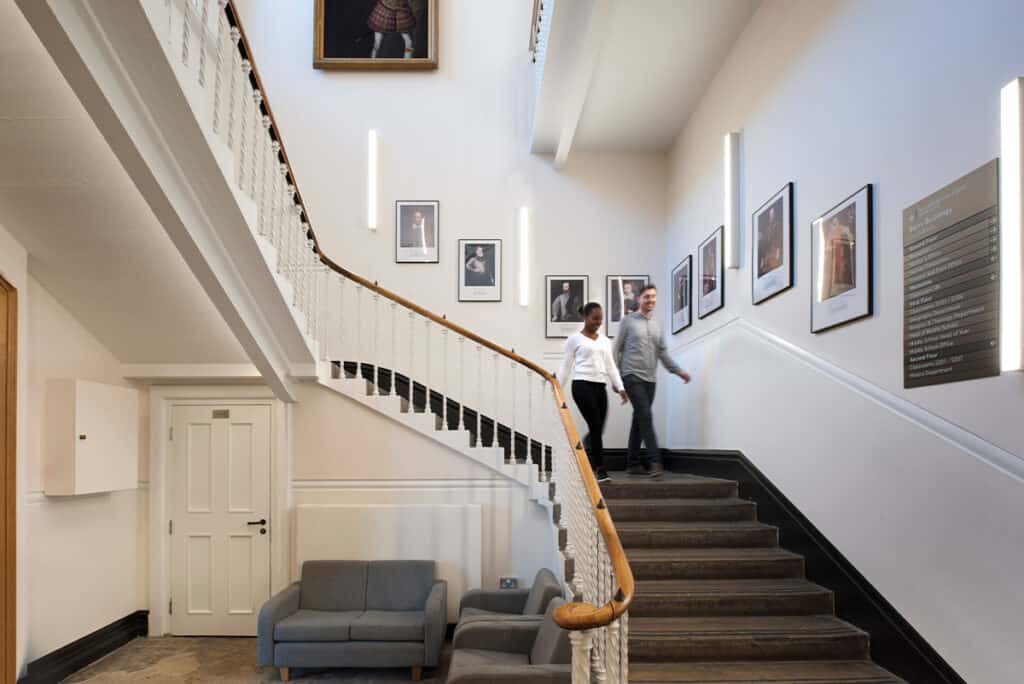 dulwich-college-5 An indoor staircase with a wooden handrail and white balusters, where two people are walking down. The wall along the staircase is decorated with framed pictures, and a plaque with text is visible on the right. A door and a small seating area with a couch are located at the bottom left corner.