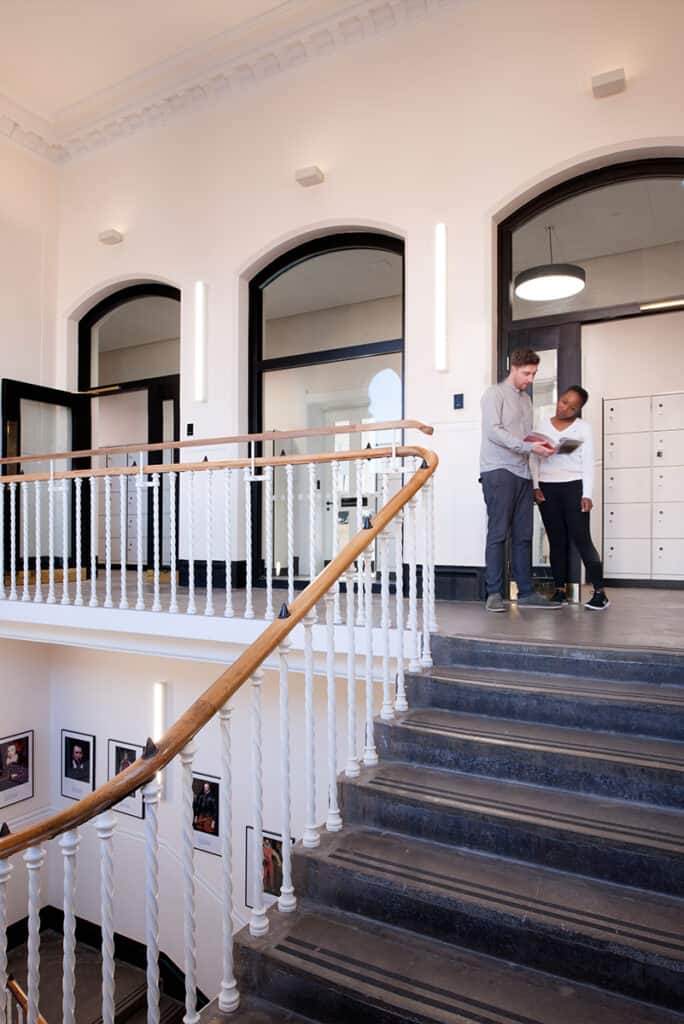 dulwich-college-6 An indoor staircase with a wooden handrail and white balusters, leading to a landing where two people stand—one holding a book or folder. The background features multiple doors, some with glass panels, a row of lockers on the right, and framed pictures on the walls.