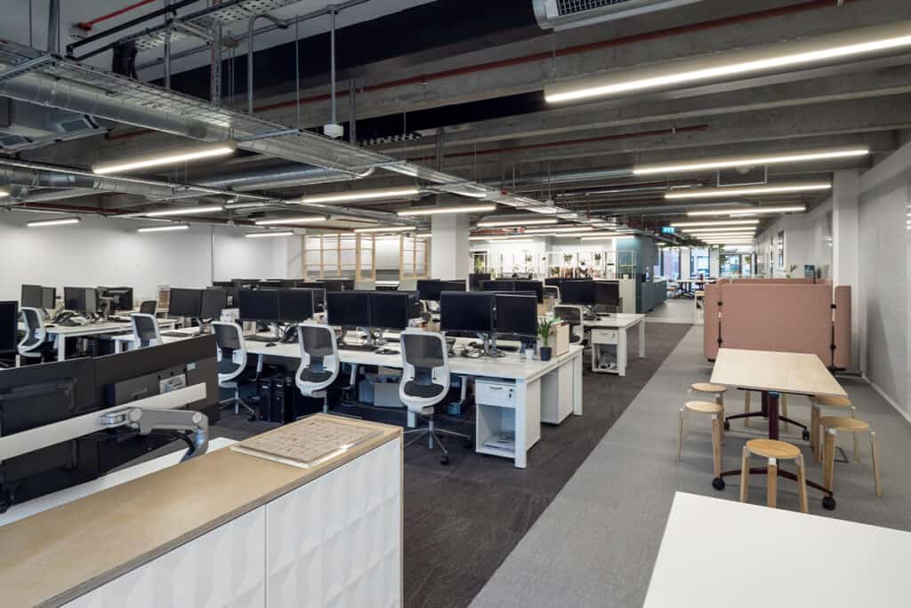 hm-architects-1 A modern open-plan office with multiple workstations, each equipped with dual monitors, ergonomic chairs, and office supplies. The space features exposed ductwork and industrial lighting. On the right, a communal table with stools adds a collaborative touch to the clean, organized layout.