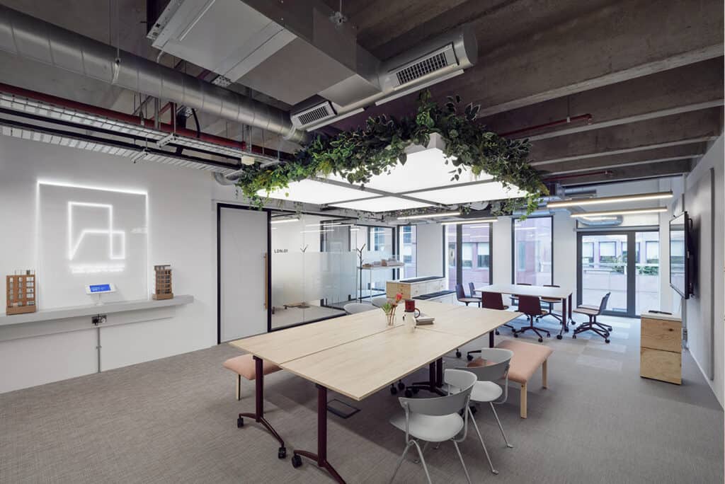 hm-architects-2 A modern open-plan office featuring a large wooden table surrounded by chairs and a bench. Ceiling lights with hanging greenery add a natural touch. In the background, glass walls and doors lead to other office areas. On the left, a lit-up logo and architectural models are displayed. The exposed ceiling with visible ducts and pipes contributes to the industrial design.
