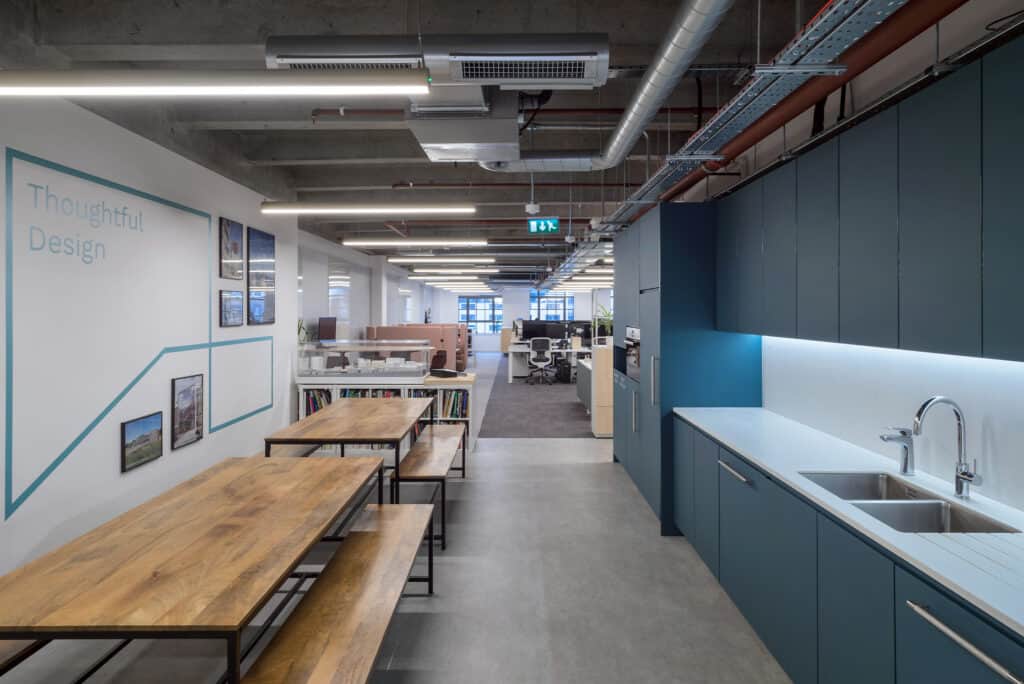hm-architects-3 A modern open-plan office space. On the left, a wall displays the text "Thoughtful Design" alongside framed pictures, with wooden tables and benches placed in front. On the right, a kitchenette features blue cabinets, a white countertop, and a sink. The background includes rows of desks and chairs, with exposed ceiling ducts and lighting fixtures overhead.