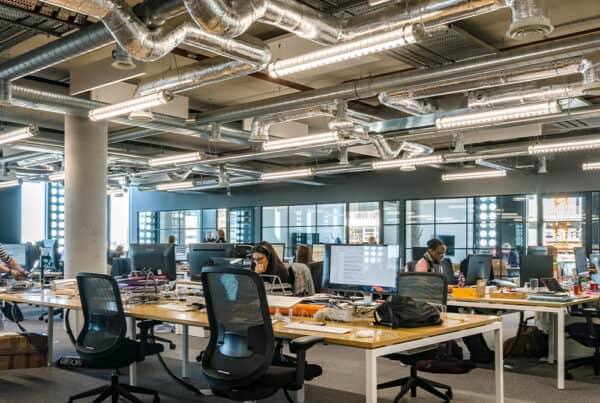 Modern open-plan office with multiple desks, computers, and office chairs. Several people are working at their stations. The space features exposed ductwork, industrial lighting, large windows, and glass partitions dividing different areas.