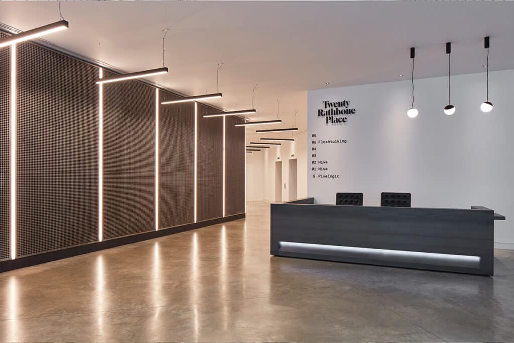 rathbone-1 Modern office reception area with a sleek dark desk, wall-mounted directory text listing office levels, vertical light strips on a patterned wall, hanging ceiling fixtures, and polished concrete flooring.