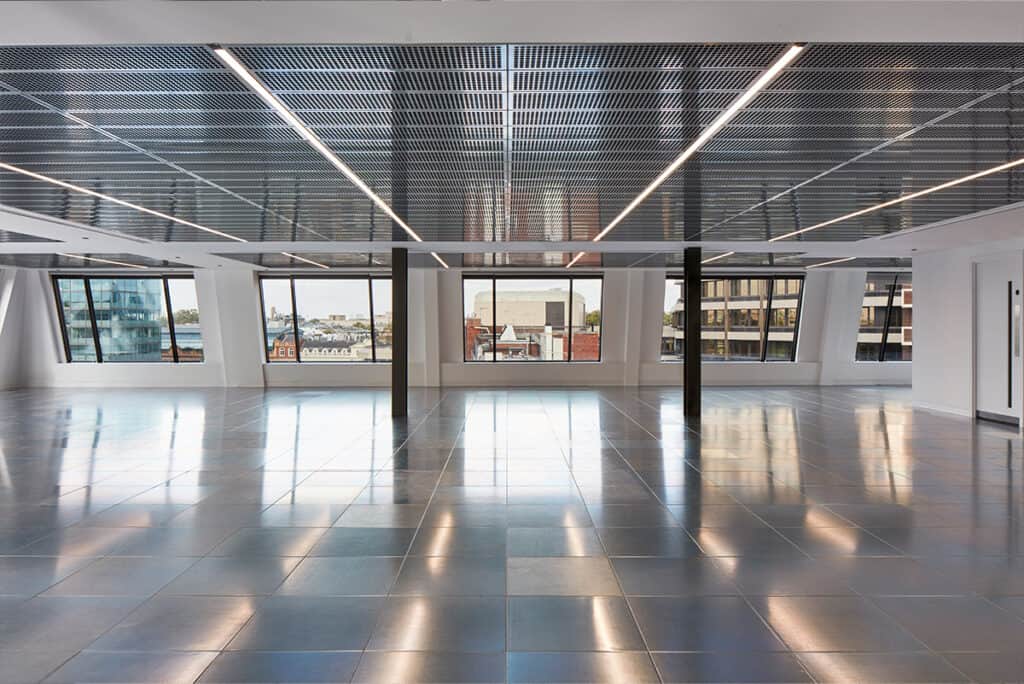 shell and core Large CAT A modern office space with a reflective tiled floor, grid-pattern ceiling with integrated lighting, and white walls. The room features two black columns and large windows that let in natural light and offer views of nearby buildings.