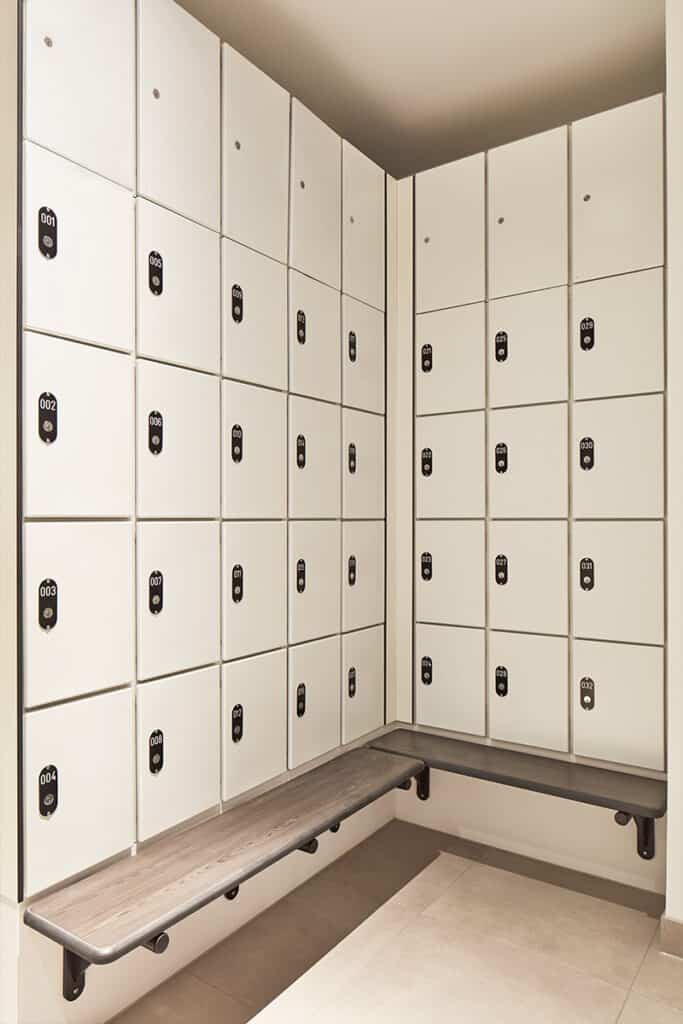 rathbone-3 Locker room corner with two walls of white lockers, each featuring black lock mechanisms labeled with numbers such as 001 to 004. A wooden bench is mounted below the lockers along both walls.