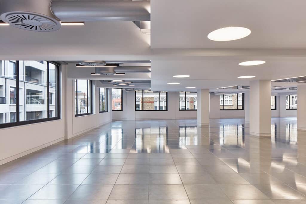 rathbone-7 Large CAT A office space with tiled flooring, white walls, and structural columns. The ceiling features circular lights and ventilation units. Numerous windows line the room, allowing natural light to fill the spacious interior.