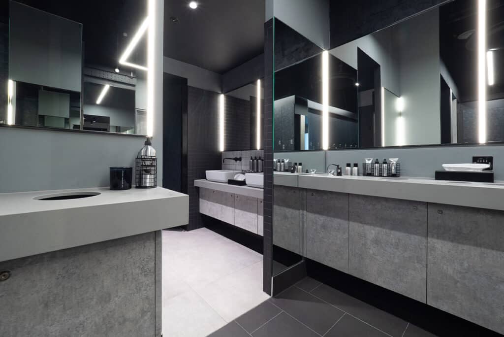 rowbots-2 Modern bathroom with a sleek, minimalist design featuring two large mirrors framed by vertical LED lights. Concrete-style countertops hold neatly arranged toiletries, and the space is styled in a gray and black color scheme for a contemporary look.