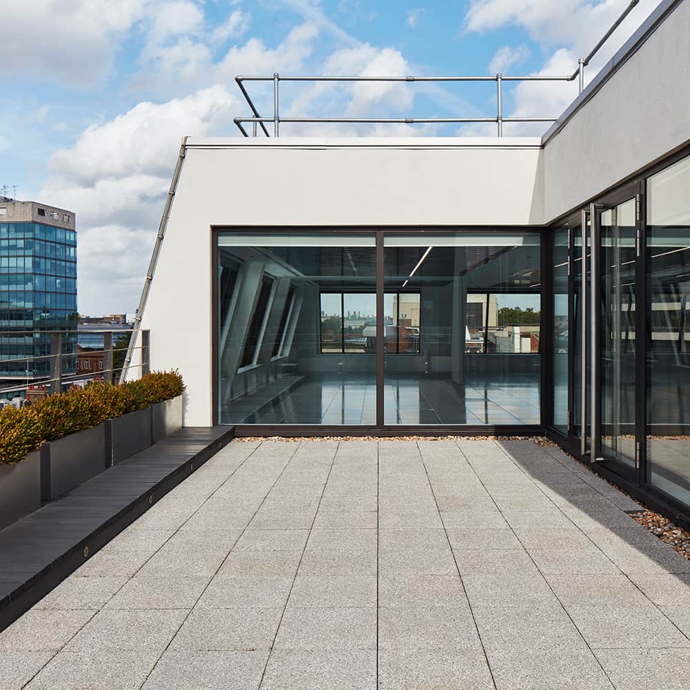 small-works-2 Modern rooftop terrace with tiled flooring, large glass windows and doors leading indoors, a planter with greenery along one side, and a view of surrounding buildings under a clear sky.