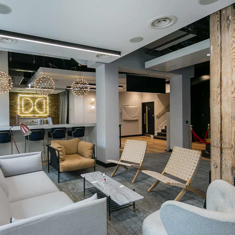 small-works-4 Modern lounge or common area with a mix of seating including a beige armchair, woven chairs, and a gray sofa. A bar area with three bar stools and spherical pendant lights sits in front of a brick wall with a neon 'DD' sign. Stairs with red velvet ropes indicate restricted access. The space features neutral tones with wood and metal accents.