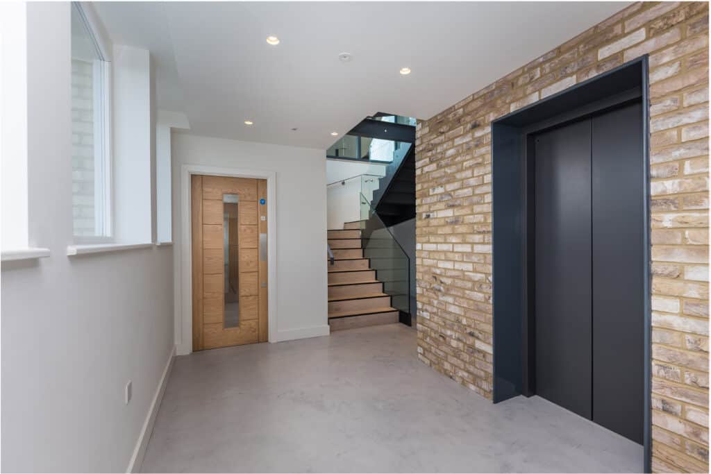 Lift Installation Modern interior space featuring a staircase with wooden steps and glass railings, an elevator with dark doors set into a brick wall, white walls, a concrete floor, and a wooden door with a vertical glass panel.