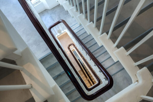 Top-down view of a multi-story staircase with grey steps, white railings, and dark brown handrails, featuring an open central area offering a view down to the lower floors.