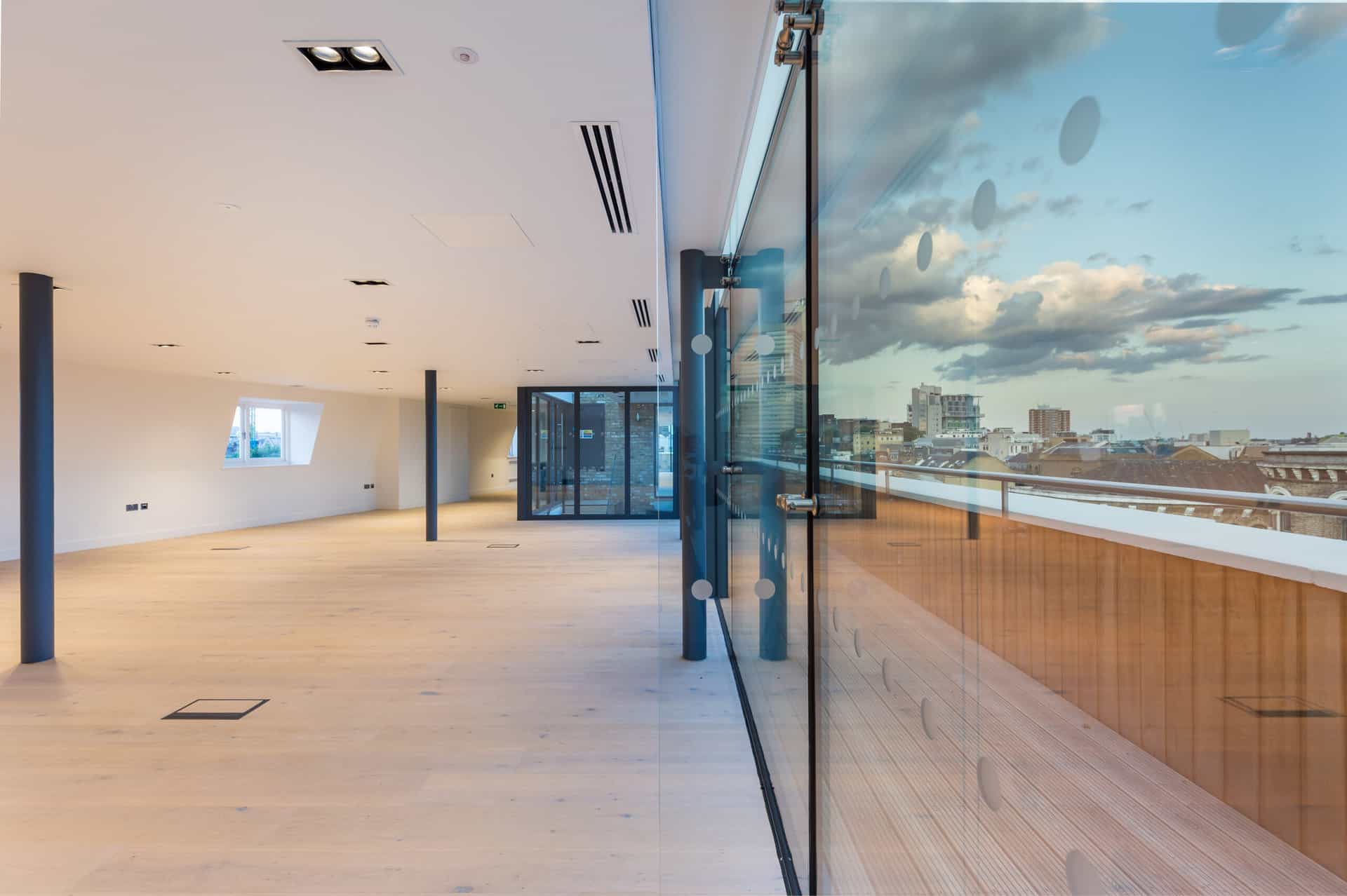 Modern empty CAT A office space with light wooden flooring, white walls, black ceiling-support columns, recessed lighting, air conditioning vents, a large glass wall reflecting outdoor buildings and sky, and glass doors at the far end.