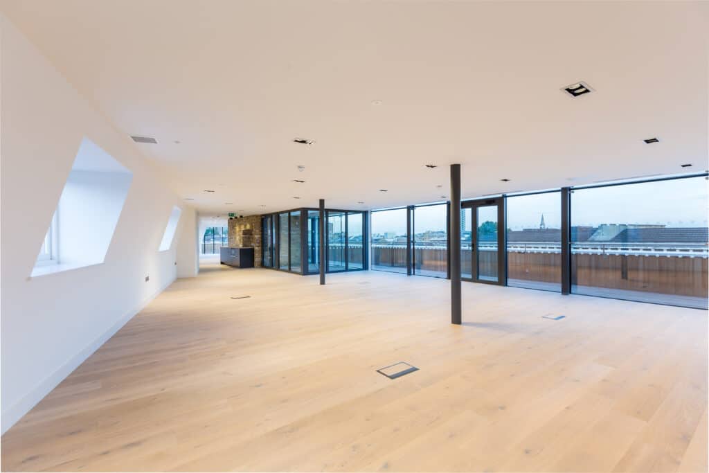 untitled-228 Spacious modern open plan office fit out with wooden flooring, white walls, recessed ceiling lighting, structural columns, large floor-to-ceiling windows offering a panoramic city view, and glass doors leading to a balcony or terrace.