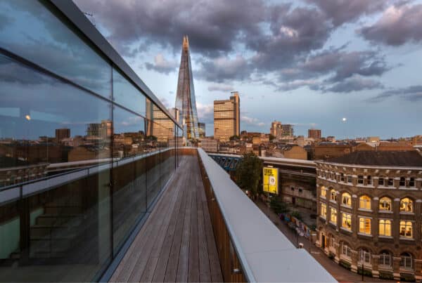 A stunning rooftop view during twilight, showcasing a skyline that includes the iconic Shard building amidst a backdrop of clouds. The foreground features a sleek glass railing and wooden decking, creating an elegant outdoor space. The cityscape below is illuminated with evening lights, adding to the vibrant atmosphere.