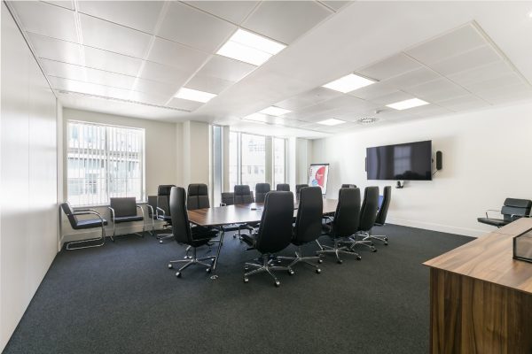 Modern conference room featuring a large rectangular table surrounded by twelve black office chairs, with additional seating along the walls, large windows with vertical blinds, a wall-mounted flat-screen TV, and a wooden cabinet in the foreground.