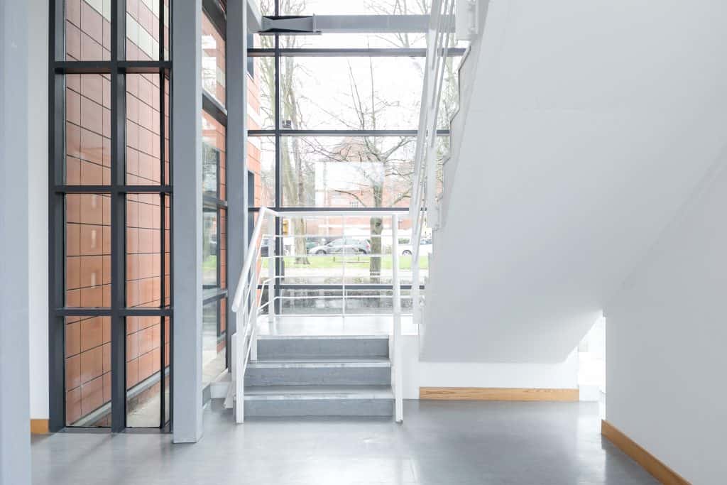 1-14 Refurbished interior stairwell featuring white railings, grey steps, and large windows that flood the space with natural light. The fit-out combines clean white walls with wooden trim, enhancing the bright and modern architectural design.