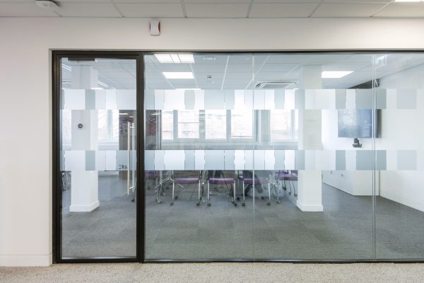 1-15 A recently refurbished conference room enclosed by full-height glass walls featuring frosted rectangular patterns for privacy and visual interest. Inside, several modern chairs with purple upholstered seats are arranged around a central meeting table, reflecting a contemporary and vibrant design choice. The room benefits from large windows that flood the space with natural light, enhancing the open and airy feel. A flat-screen TV is mounted on the right-hand wall, integrated for presentations and video conferencing. The flooring is fully carpeted, contributing to acoustic comfort, while the ceiling includes recessed LED lighting and integrated air conditioning vents, indicating a focus on energy efficiency and occupant comfort. The refurbishment emphasizes transparency, collaboration, and modern workplace functionality.