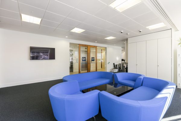 Modern office waiting area featuring a circular arrangement of blue couches around a black glass coffee table, with a wall-mounted flat-screen TV, wooden double doors with glass panels, and an elevator visible in the background. Recessed ceiling lights and a large white cabinet complete the space.