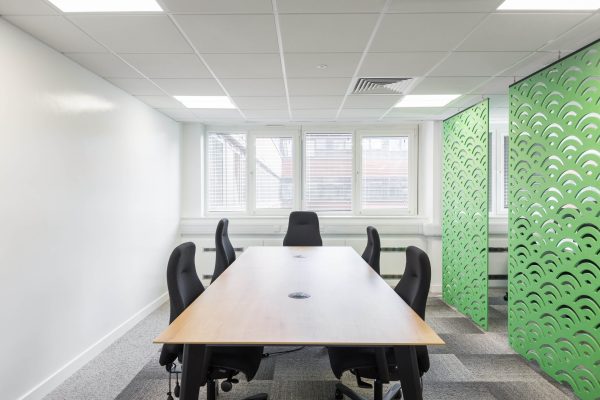1-22 A recently refurbished conference room featuring a sleek rectangular wooden table surrounded by six black ergonomic office chairs. The space is defined by clean white walls and a white ceiling fitted with recessed LED lighting, enhancing brightness and energy efficiency. Three large windows line the far wall, fitted with adjustable blinds to control natural light and maintain privacy. A green decorative partition on the right adds a pop of color and visual interest, potentially serving as an acoustic or biophilic design element. The overall refurbishment reflects a minimalist, modern aesthetic focused on functionality, comfort, and a professional atmosphere.