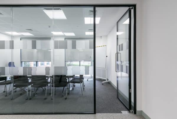 Refurbished conference room featuring glass walls with frosted privacy patterns, a long table with wheeled chairs, and a mobile whiteboard. The fit-out includes recessed ceiling lighting, dark carpeting, and partially shaded windows for a clean, functional meeting space.
