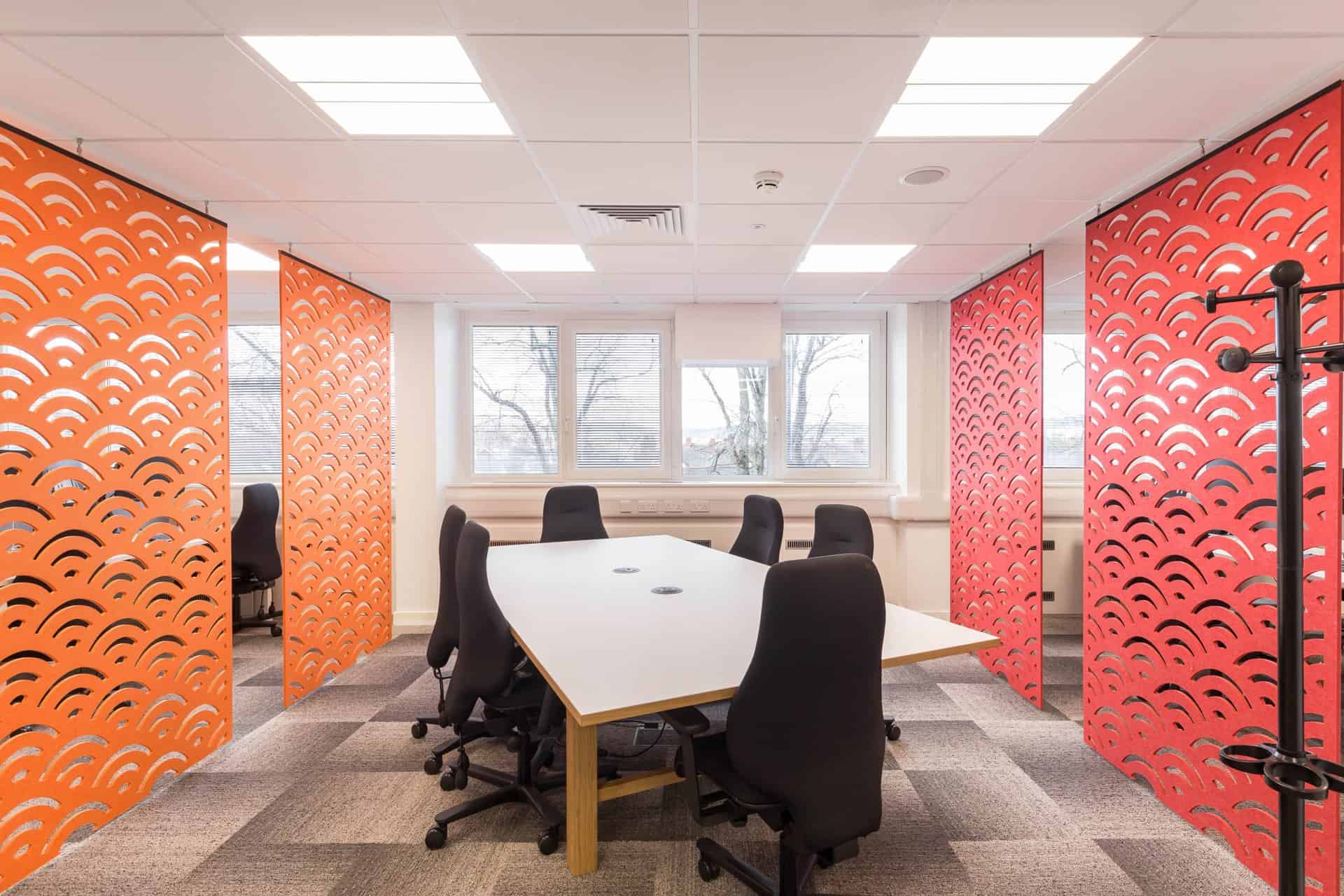 A newly refurbished modern office meeting area featuring a large rectangular table with rounded edges, surrounded by five black ergonomic chairs on castors. The flooring has been updated with grey and white carpet tiles laid in a checkered pattern, offering a clean and contemporary look. The ceiling includes recessed LED lighting panels, contributing to energy efficiency and a bright workspace. Three large windows at the far end of the room allow ample natural light, enhanced by partially raised horizontal blinds. On both sides of the table, bold orange-red partition panels with artistic wave-like cutouts have been installed, adding visual interest and acoustic separation. A sleek black coat rack is positioned near the right partition, complementing the modern aesthetic. The overall refurbishment emphasizes functionality, comfort, and a vibrant yet professional atmosphere.